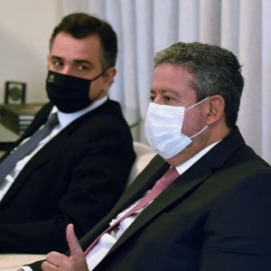 Foto de Arthur Lira, presidente da Câmara dos Deputados, e Rodrigo Pacheco, presidente do Senado. Lira está em primeiro plano e Pacheco está sentado ao lado, olhando para ele.