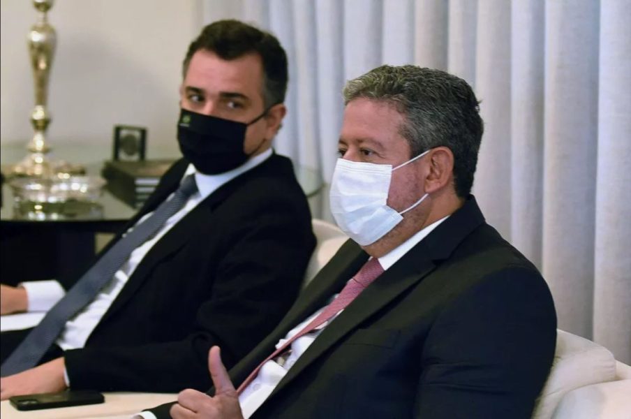 Foto de Arthur Lira, presidente da Câmara dos Deputados, e Rodrigo Pacheco, presidente do Senado. Lira está em primeiro plano e Pacheco está sentado ao lado, olhando para ele.