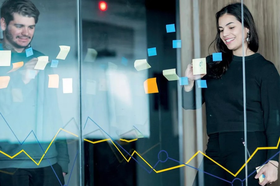 Temporada de balanços, Lucro líquido da Yduqs (YDUQ3) cai 80% no 3º tri com aumento de despesas financeiras, Mulher de pé mexe em post-its colados sobre vidro