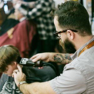 Foto de um cabeleireiro cortando o cabelo de uma criança. Ele usa a máquina, tem óculos e braço tatuado.