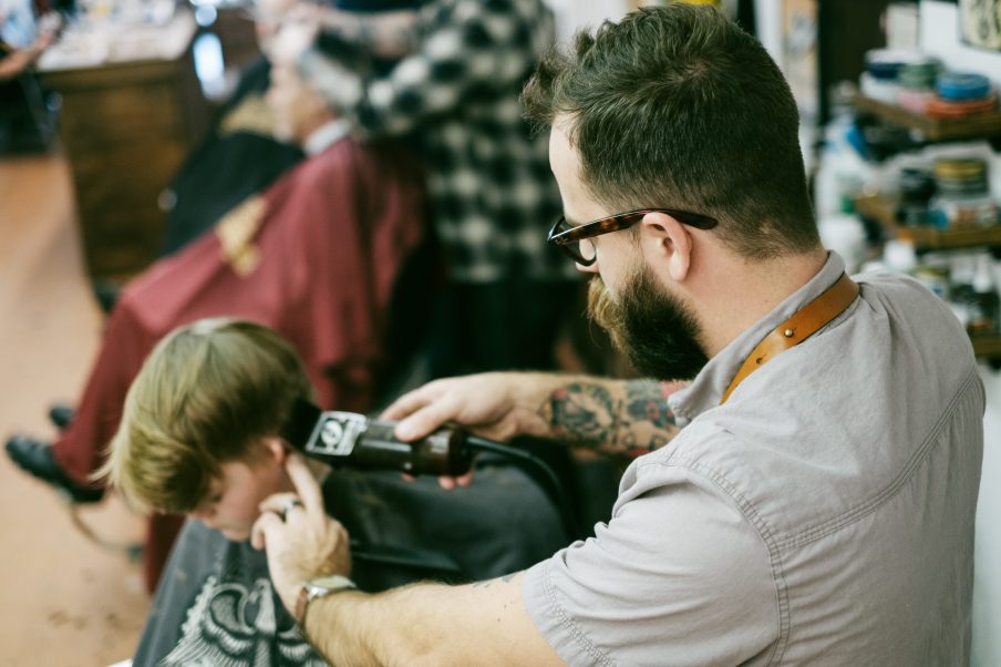 Foto de um cabeleireiro cortando o cabelo de uma criança. Ele usa a máquina, tem óculos e braço tatuado.