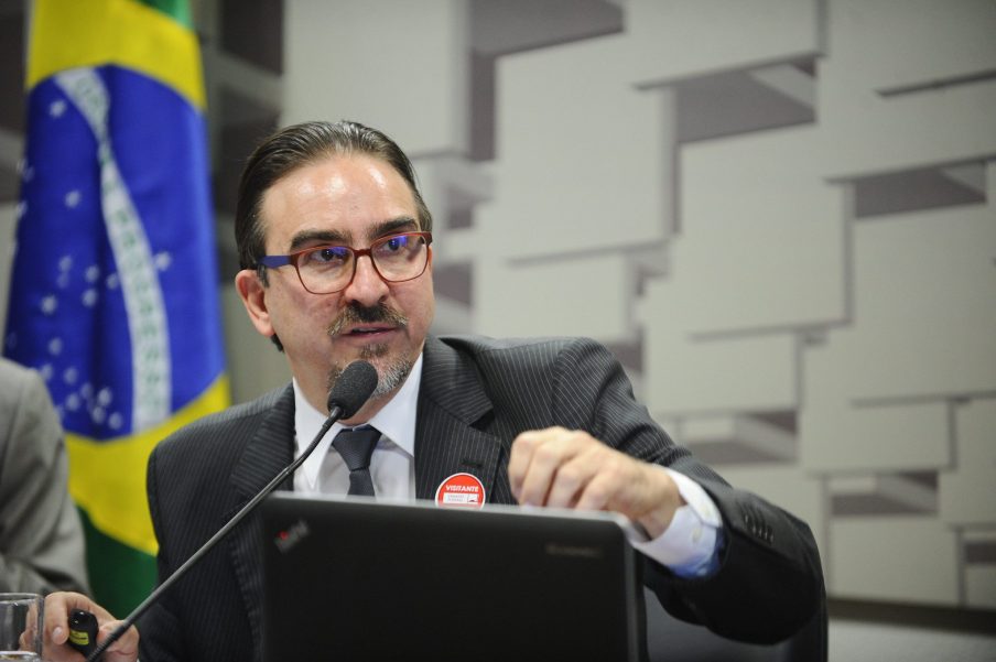 O economista Bernard Appy fala a um microfone com a bandeira do Brasil no fundo