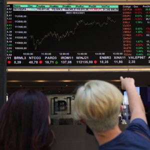 Foto de duas pessoas apontando para um painel com gráfico de linha sobre a bolsa de valores. Matéria detalha o movimento do Ibovespa hoje (17/05/2024)