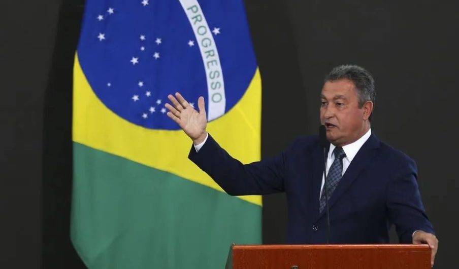 Foto do ministro Rui Costa discursando com a bandeira do Brasil ao fundo