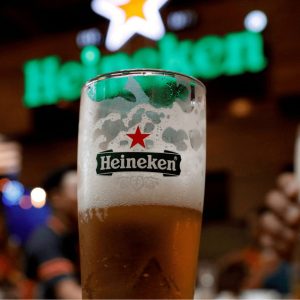 Foto de um copo de cerveja da Heineken com cerveja preenchendo a metade do copo com espuma no topo. Ao fundo está o logo da marca