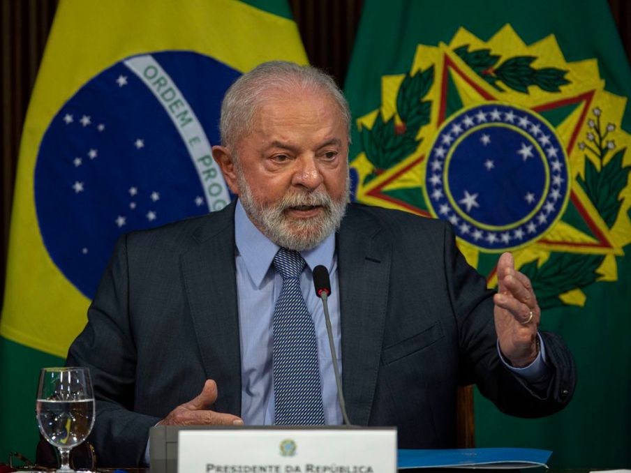 Foto de Luiz Inácio Lula da Silva, presidente da República. Ele usa um terno preto, gravata com estampas e camisa azul clara. Fala em um microfone com as bandeiras do Brasil e do governo federal ao fundo