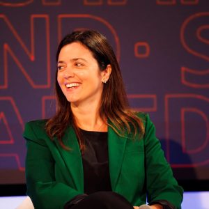 Foto da analista de fundos Luciana Seabra. Ela é mulher, veste uma camisa preta e um blazer verde, e está olhando para o lado e sorrindo