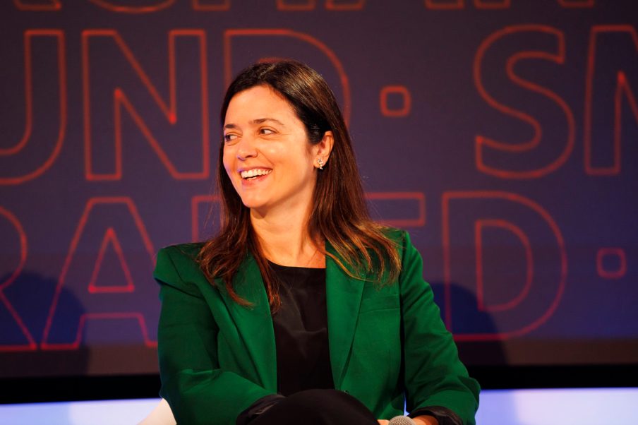 Foto da analista de fundos Luciana Seabra. Ela é mulher, veste uma camisa preta e um blazer verde, e está olhando para o lado e sorrindo