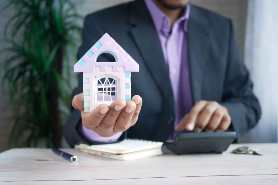 Foto para a matéria sobre quanto rendem R$ 5 mil em fundos imobiliários em que aparece um homem segurando uma maquete de uma casa e com a outra mão fazendo cálculos em uma calculadora.