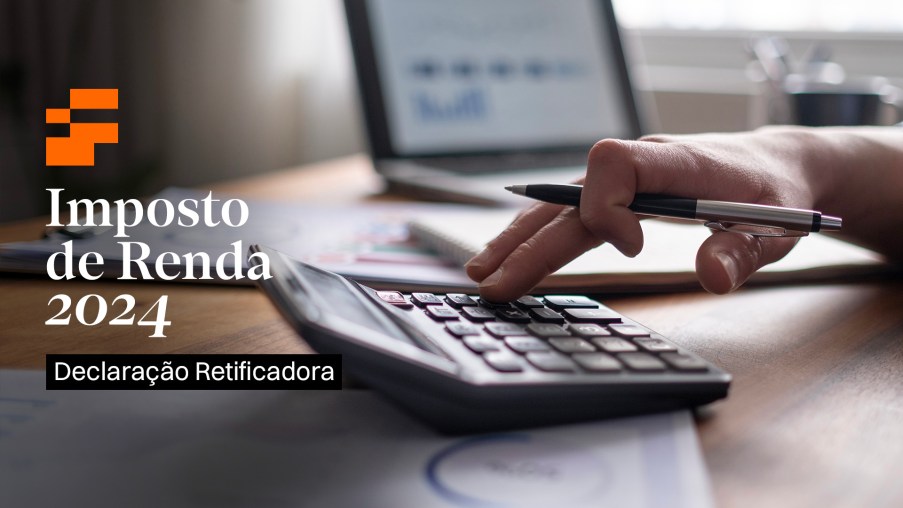 Foto mostra apenas objetos e uma mão. Foco está em uma calculadora sendo operada pela mão de uma pessoa e ao fundo um computador. Os objetos estão sob uma mesa de madeira. Tema da matéria é Declaração de Imposto de Renda 2024