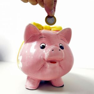 Foto mostra mão colocando moeda de R$ 1 em um cofrinho rosa para ilustrar reportagem sobre quanto rendem R$ 100 mil no Tesouro Selic