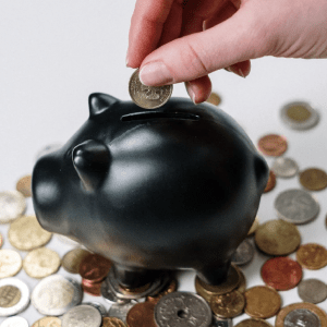 Porquinho preto com uma mão inserido moedas. Abaixo dele, moedas espalhadas