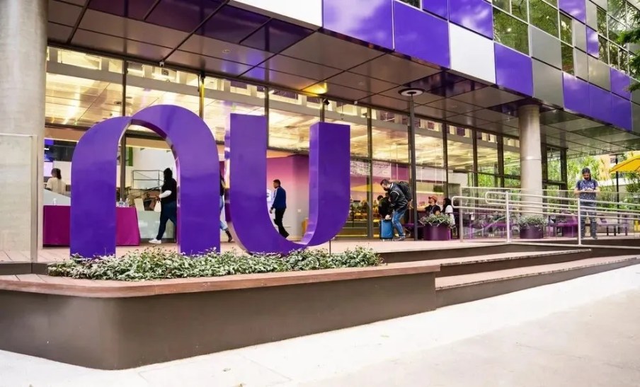 Foto da fachada do escritório do Nubank. Há um letreiro escrito "Nu" em roxo em frente a um prédio também com detalhes da mesma cor.