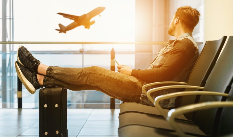 Imagem sobre destinos brasileiros baratos em que aparece um homem sentado em um aeroporto com as pernas esticadas em cima da mala olhando um avião subir.