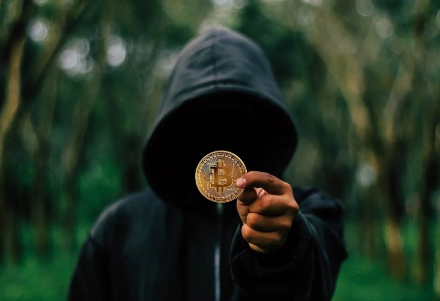 Imagem de uma pessoa com um capuz, com uma blusa preta com capuz. O rosto está tampado por uma sombra. Nas mãos, está segurando uma moeda com o símbolo do bitcoin.