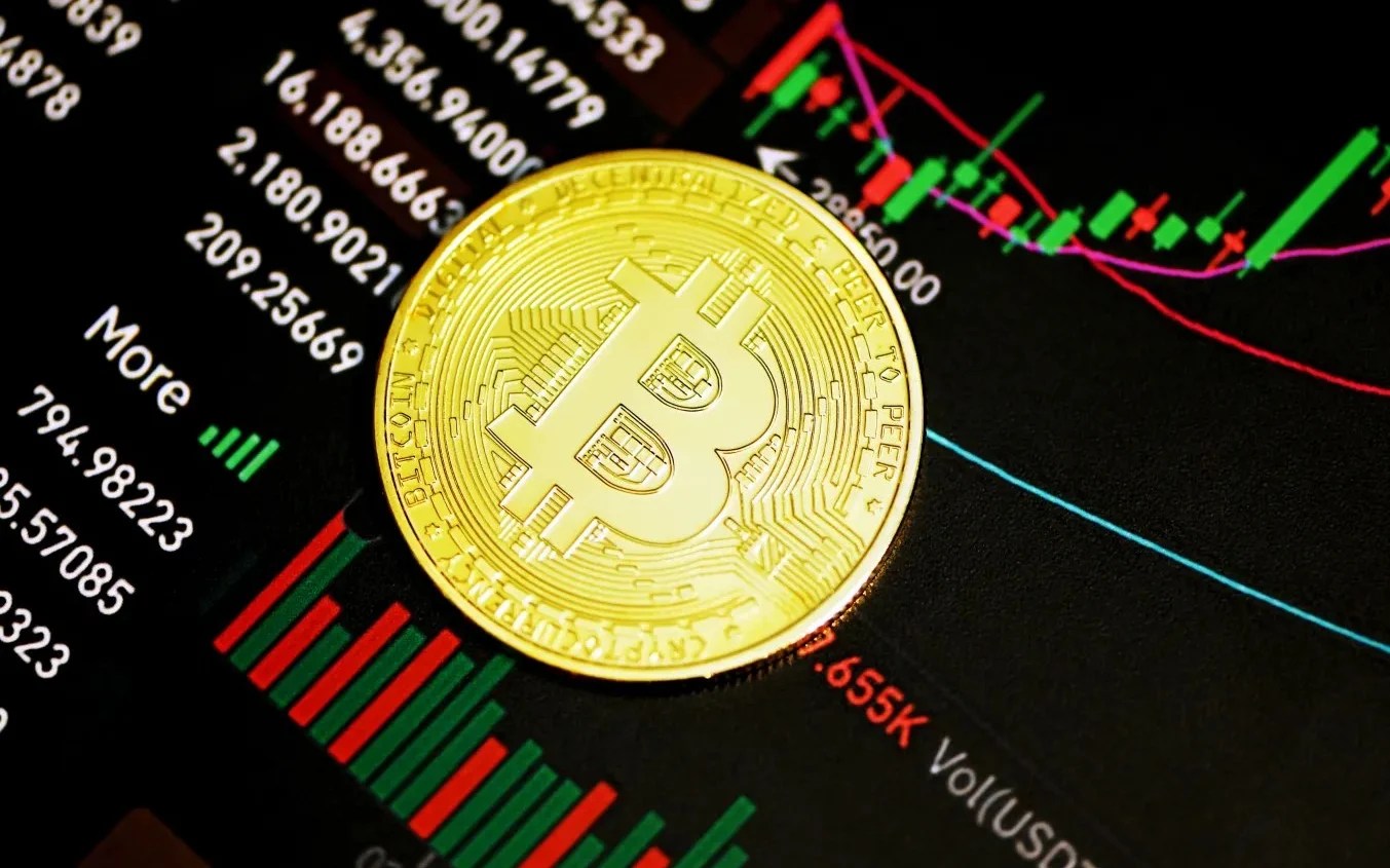 Foto de um bitcoin com cotações de ativos financeiros ao fundo, com gráfico de barras. A matéria explica a alta das criptomoedas em 2023.