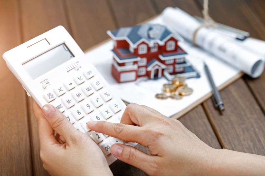 Imagem para a matéria sobre como investir em imóvel em que aparecem duas mãos segurando uma calculadora e ao fundo uma maquete de uma casa com uma pilha de moedas na frente.