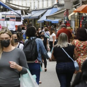 Foto de rua movimentada por pessoas com lojas ao redor. A matéria fala sobre investimentos em ações do setor de varejo na bolsa de valores.