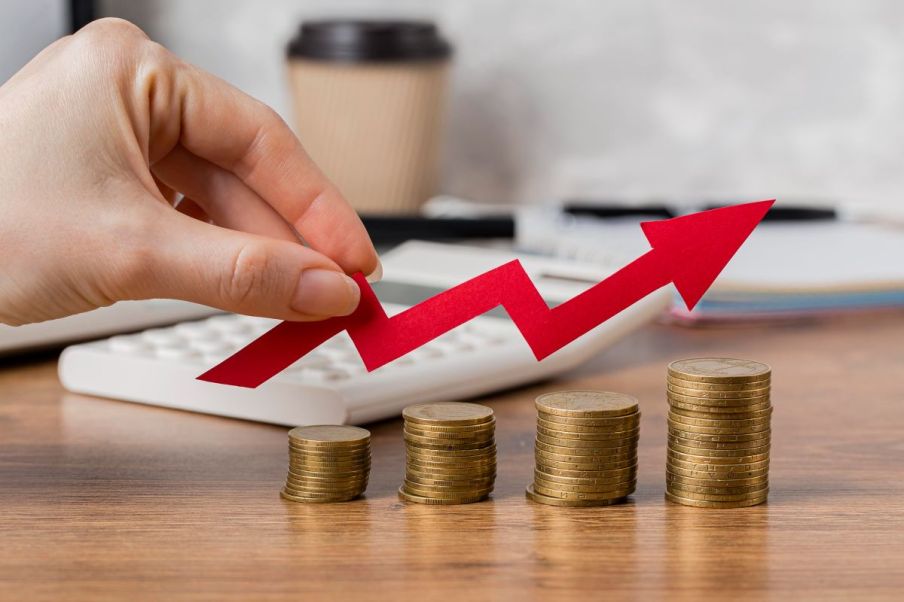 Imagem para a matéria sobre investir em debêntures em que aparece uma pilha de moedas crescentes, uma mão segura uma seta que indica subida. Ao fundo uma calculadora e um copo de café.