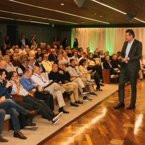 Imagem para matéria do Itaú Unibanco (ITUB4) em que aparece um homem branco apresentando o evento com várias pessoas, entre homens e mulheres, na platéia.