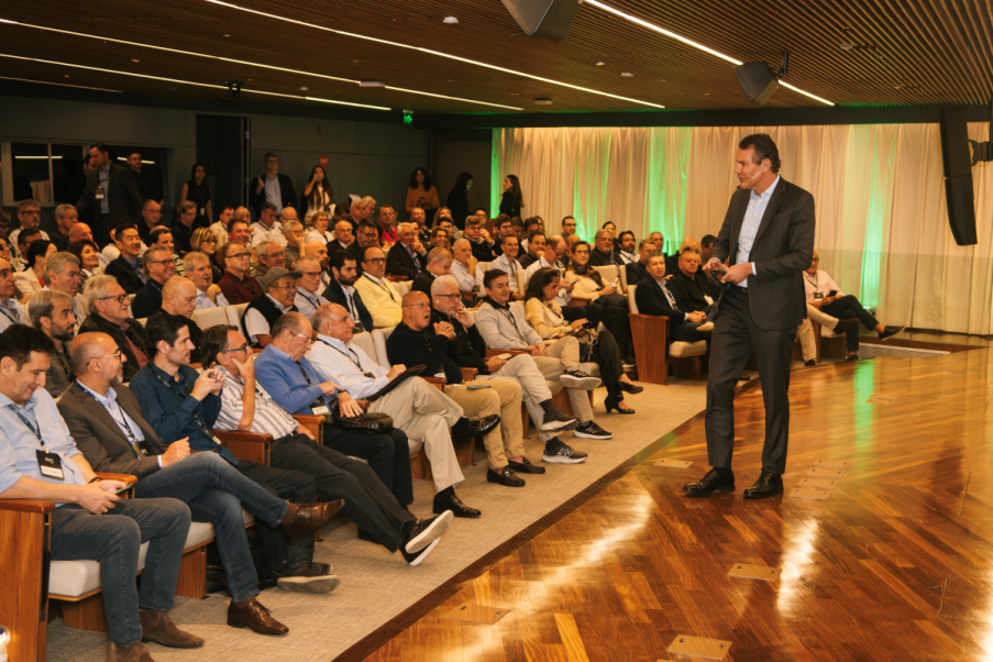 Imagem para matéria do Itaú Unibanco (ITUB4) em que aparece um homem branco apresentando o evento com várias pessoas, entre homens e mulheres, na platéia.