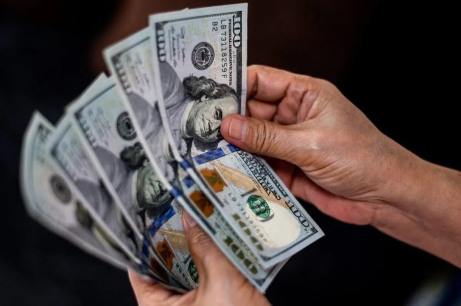 Foto de mão segurando notas de dólar americano. A matéria explica se p dólar pode chegar à cotação de 6 reais após alta da moeda.