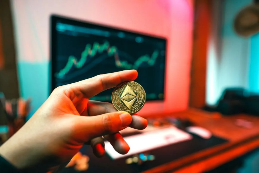 Na imagem, uma mão segura uma moeda de ethereum com uma computador ao fundo.