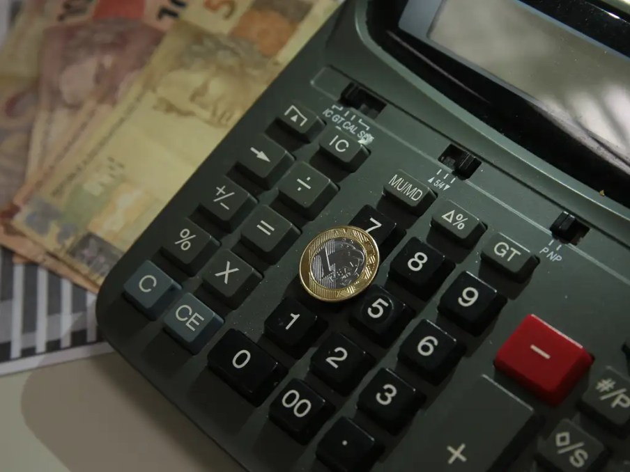 Imagem de calculadora, moeda e notas de real para ilustrar reportagem sobre onde investir R$ 1 mil