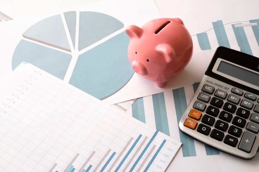 Imagem para matéria sobre CDB pós fixado em que aparecem vários papéis com gráficos, uma calculadora em cima dos papeis e um cofrinho em formato de porco.