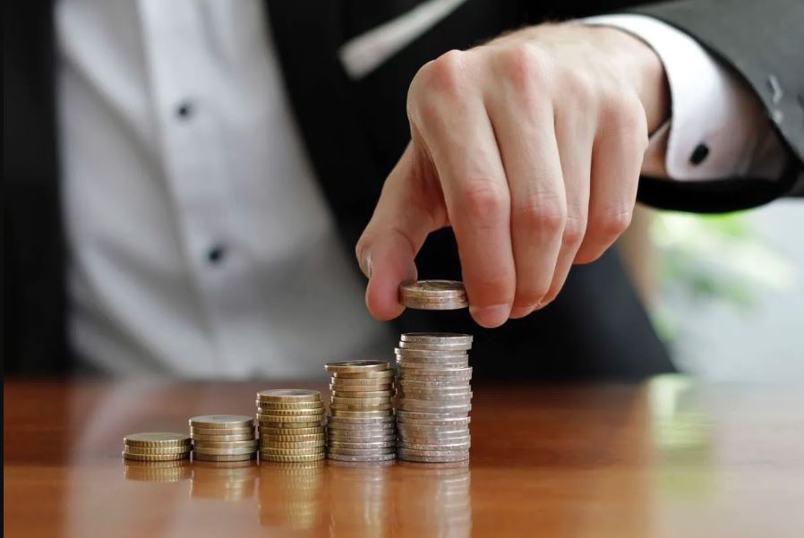 Foto de uma mão masculina colocando moedas em uma pilha. Ao lado dessa, há outras pilhas menores.