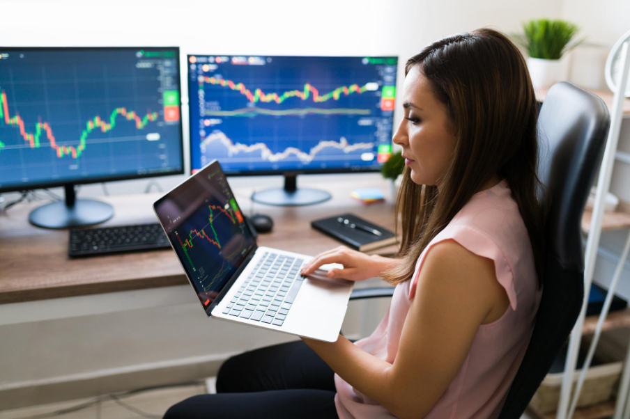 Imagem de uma mulher branca segurando um notebook olhando gráficos de investimentos. Ao fundo, duas telas de computador também com gráficos de investimentos