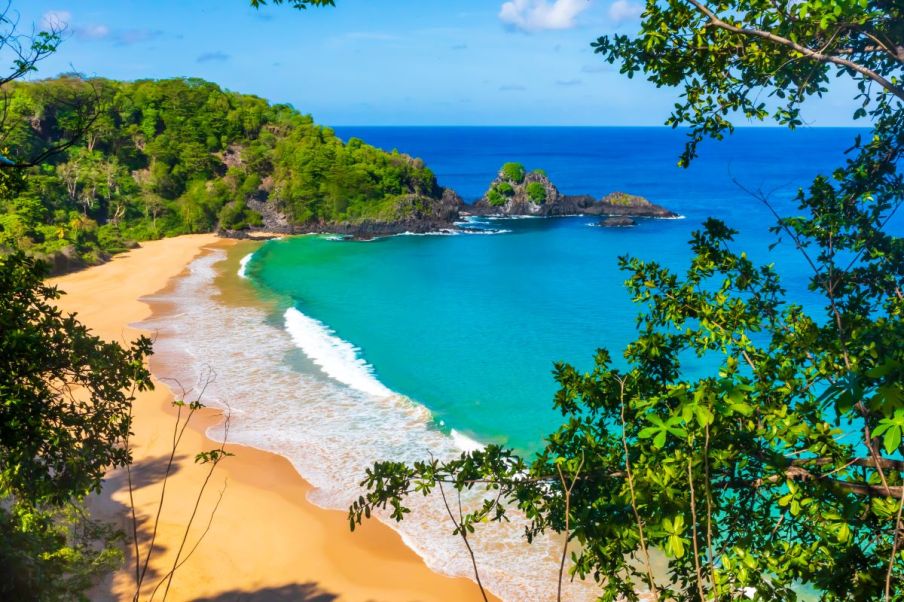 Imagem áerea de uma parte de Fernando de Noronha, a Baía do Sancho: uma praia com águas cristalinas e muitas árvores em volta.