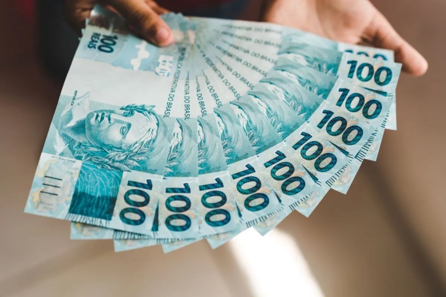 Imagem de uma mão segurando várias notas de 100 reais.