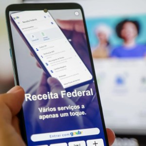 Foto de uma mão segurando um celular. Na tela, há um escrito "Receita Federal" com um fundo azul.
